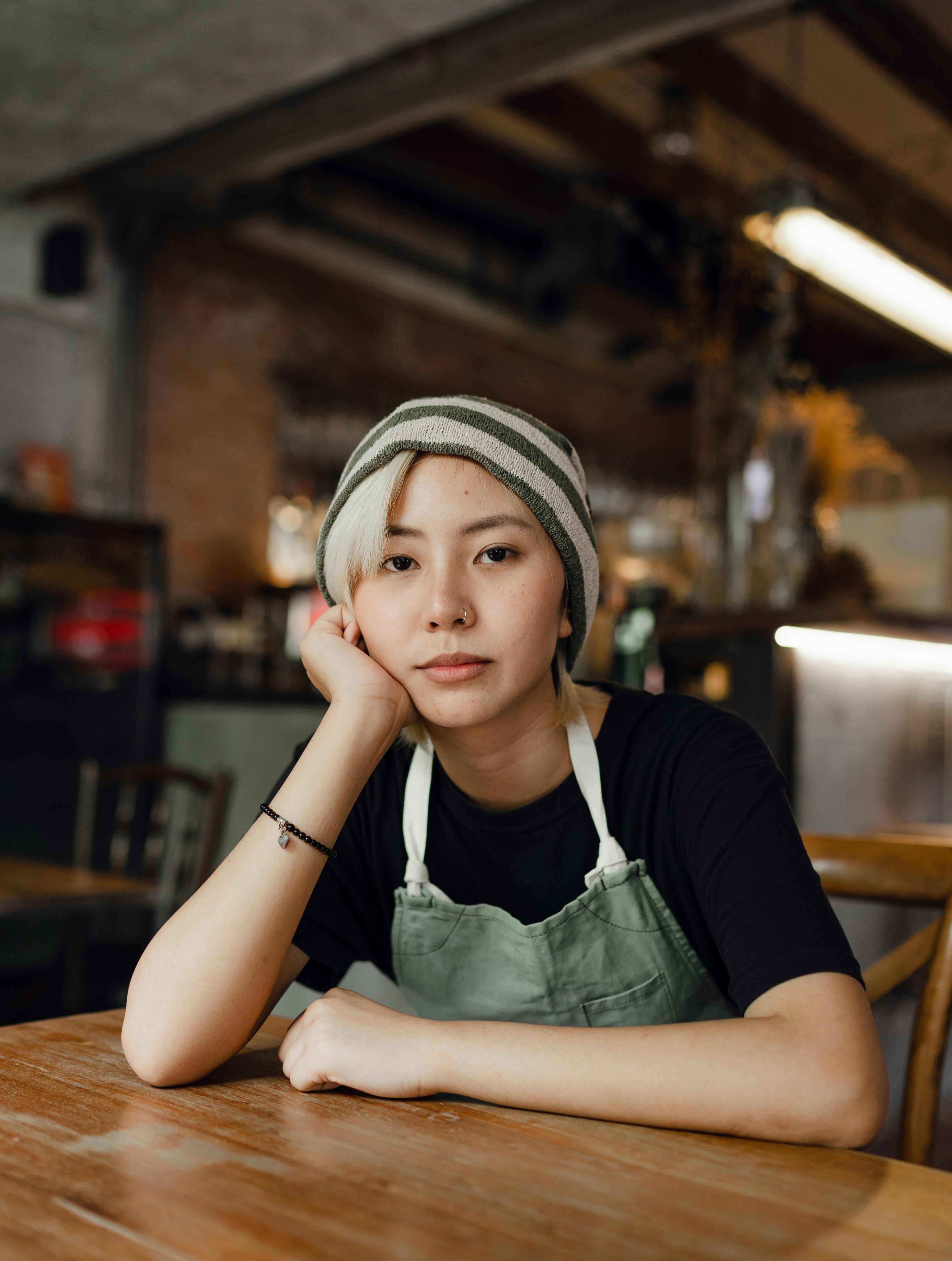 A woman wearing an apron sits at a coffee shop table with her head tilted into her palm.  A tuft of blonde hair escapes from underneath a striped beanie.