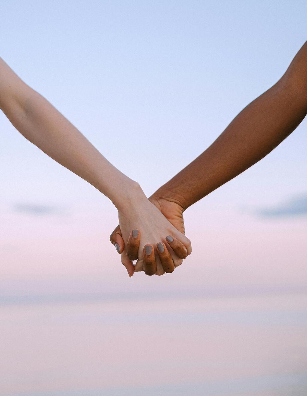 Two, multi-racial hands are clasped lovingly together with fingers interwoven.  The background is heavily blurred beach.