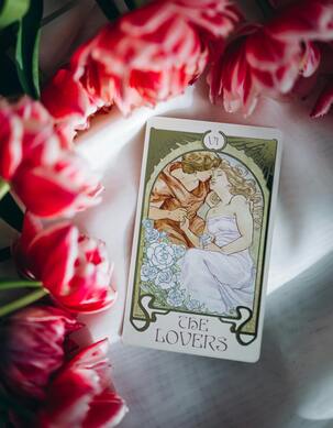 The Lovers tarot card on a rippled white fabric background, surrounded by pink flowers.